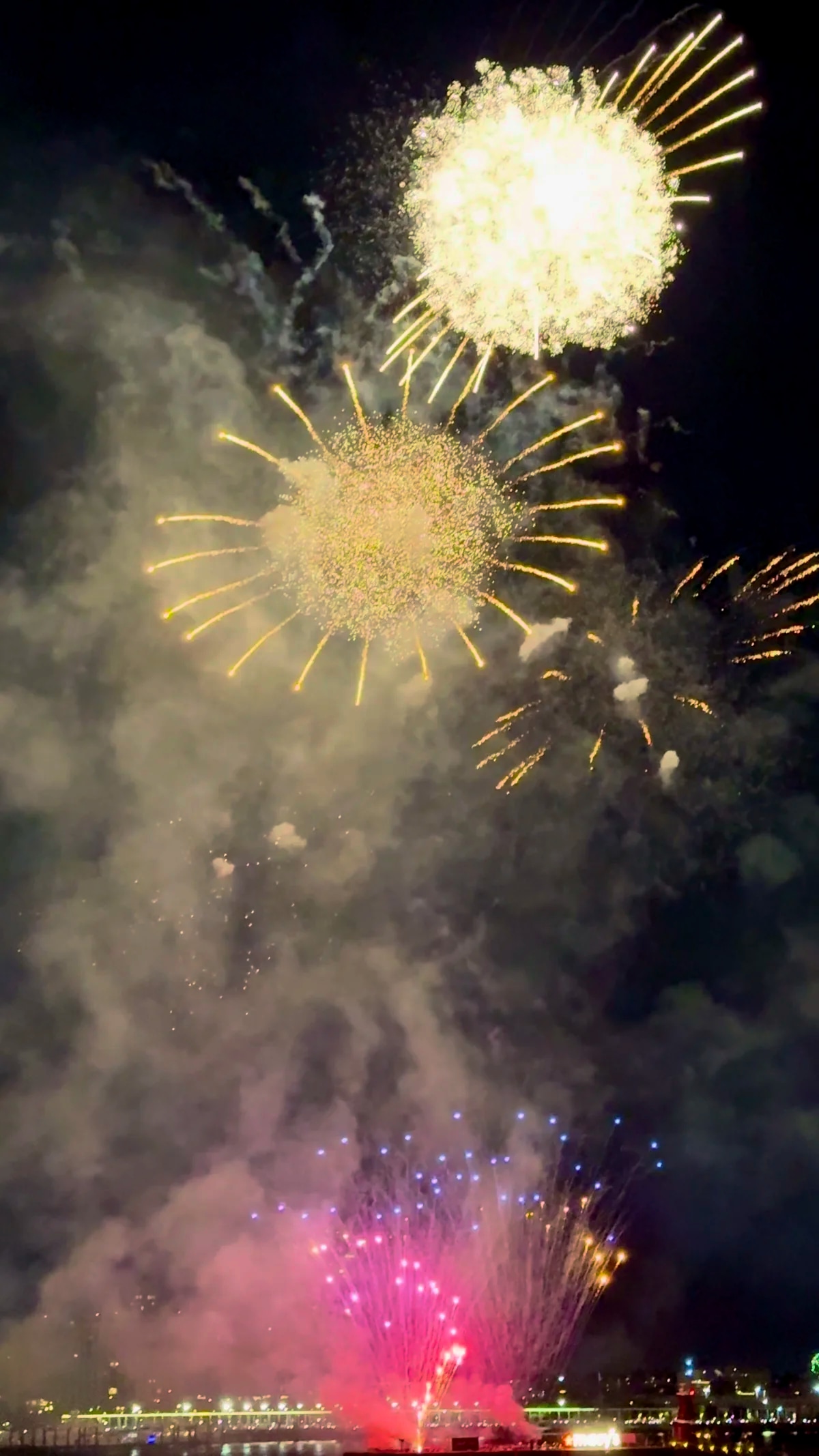 Gold and pink fireworks bursting through smoke above the water at night.